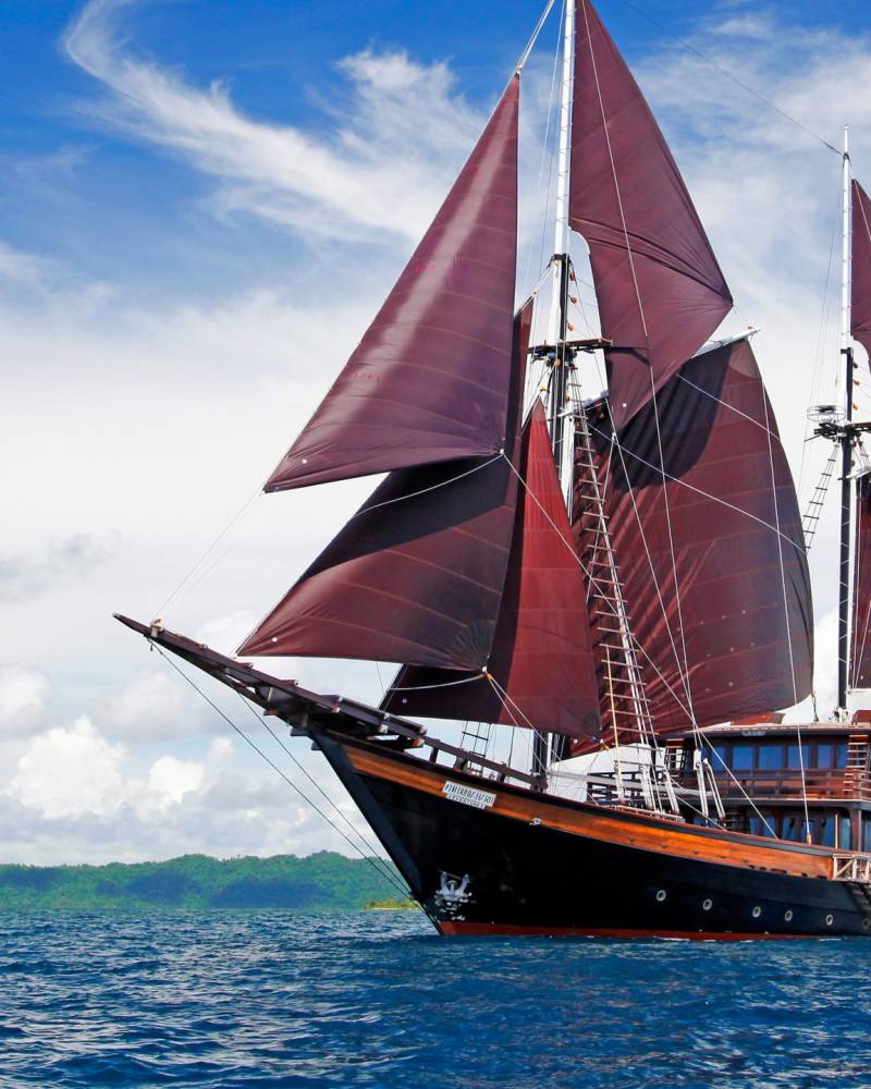 A majestic wooden schooner with deep red sails glides across the vibrant blue ocean near a lush green island.