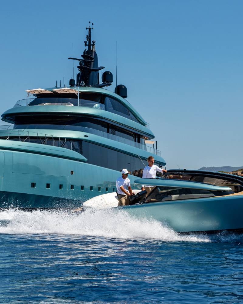 A sleek turquoise yacht glides through the sparkling blue waters near a rocky coastline under a clear sky.