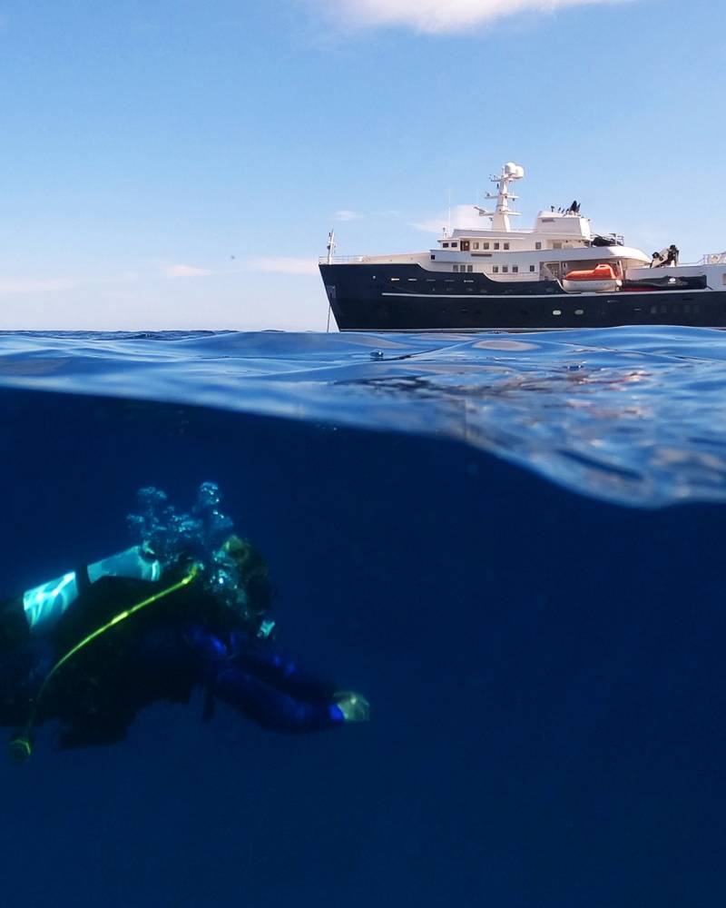 A scuba diver explores the deep blue sea beneath a luxury yacht under a clear sky.