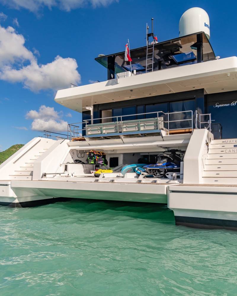 A sleek white catamaran yacht with water toys stored on its deck floats serenely in turquoise waters under a bright blue sky.