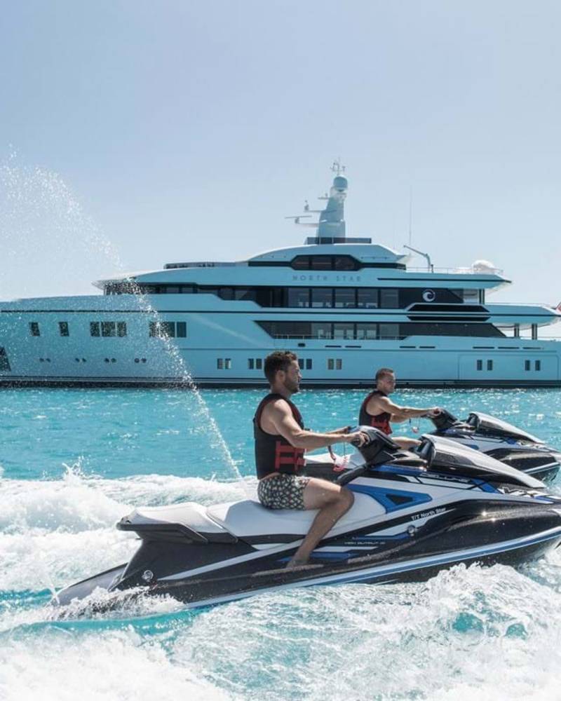 Two men on jet skis race across turquoise waters with a large yacht named "North Star" anchored in the background.