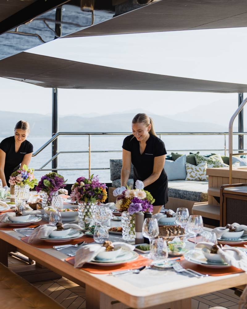 Two attendants meticulously set a luxurious outdoor dining table on a yacht overlooking a serene seascape.