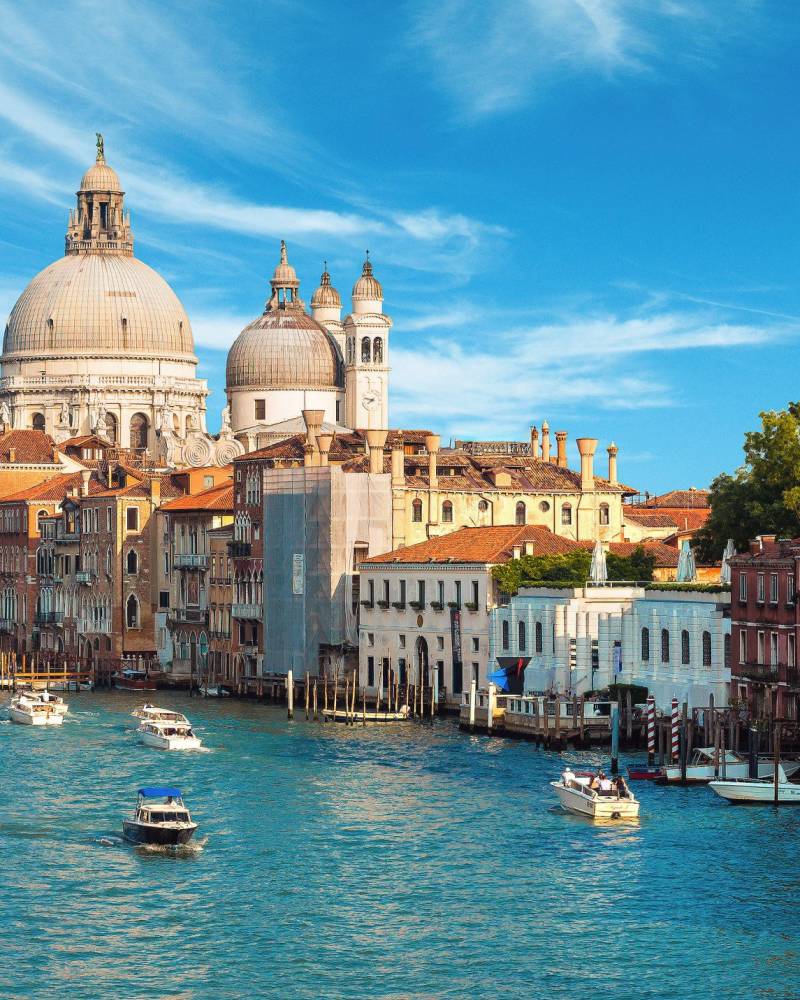 A gondola glides along the Grand Canal in Venice, with historic buildings and domes lining the waterway under a bright blue sky.