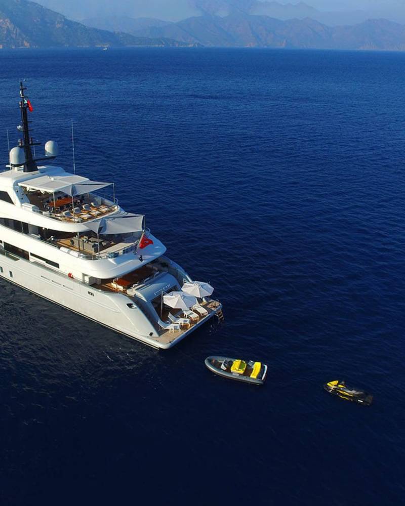 A luxurious white yacht cruises through deep blue waters near a mountainous coastline, accompanied by a tender and jet skis.