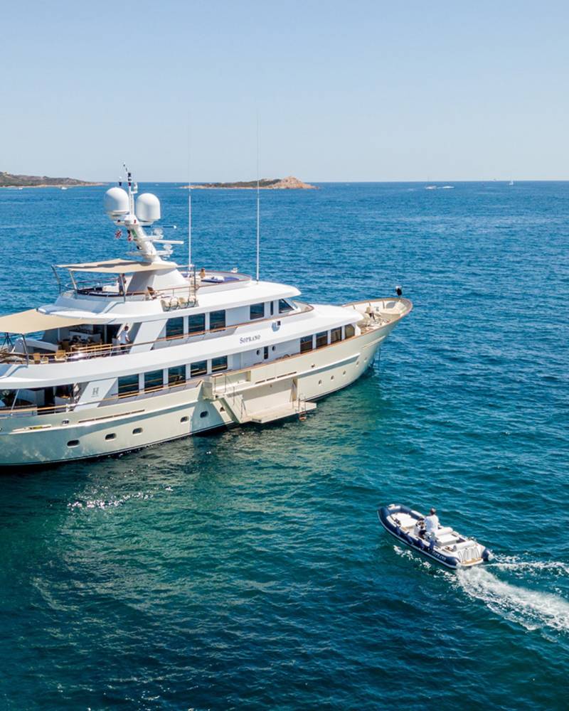 A luxurious yacht named Soprano glides through the clear blue waters near a distant, sun-drenched island.