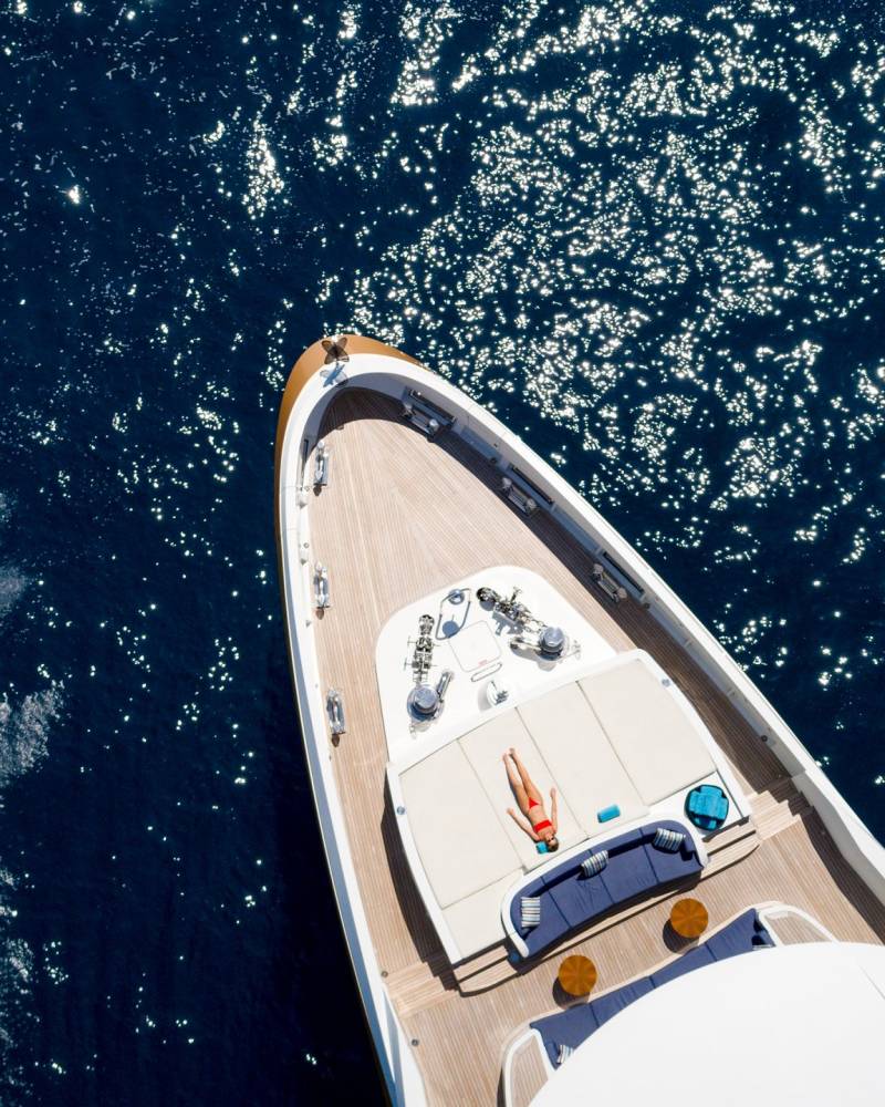 A jet ski speeds through the sparkling blue ocean near a luxurious yacht where a woman sunbathes on deck.