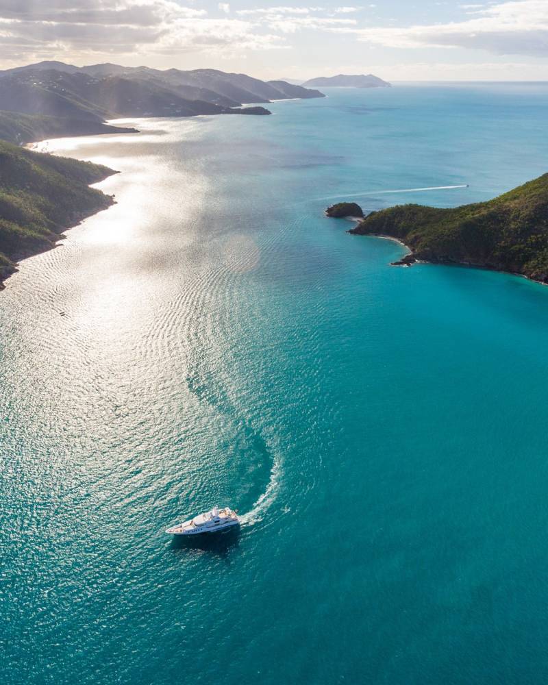 A sleek white yacht carves a white wake through the impossibly blue waters of a tropical bay.