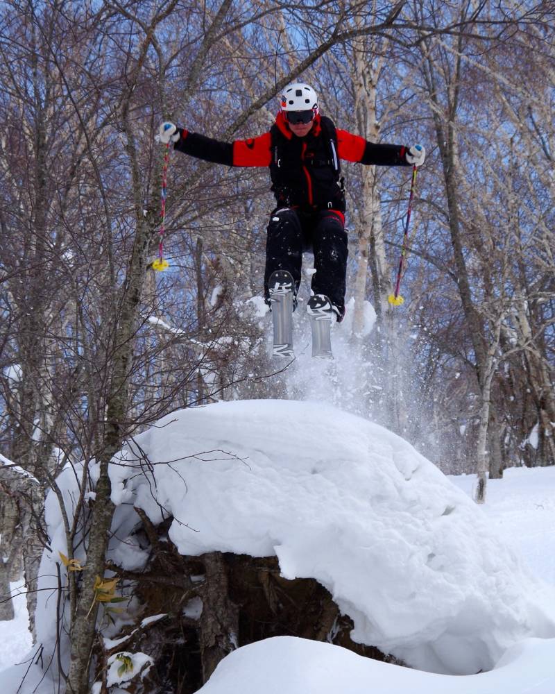 A skier soars through the air over a snowy mound amidst a forest of bare trees.