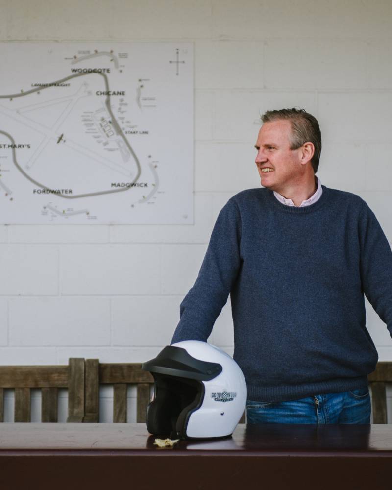 A man in a blue sweater stands by a table with a racing helmet, a map of the Goodwood Motor Circuit behind him.