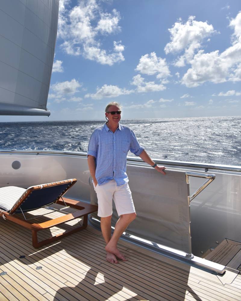 A man in a blue shirt and white shorts smiles while standing on the deck of a yacht under a bright blue sky.