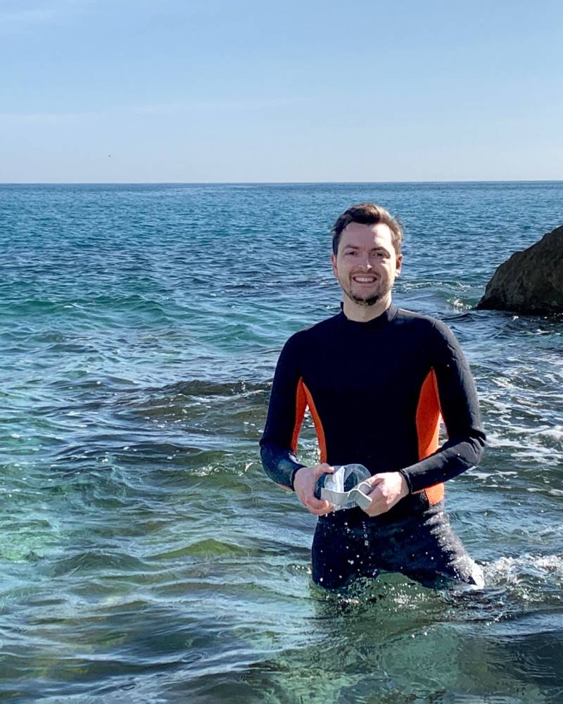 A smiling man in a wetsuit stands in the clear, turquoise waters of the Mediterranean Sea, holding a snorkel mask.