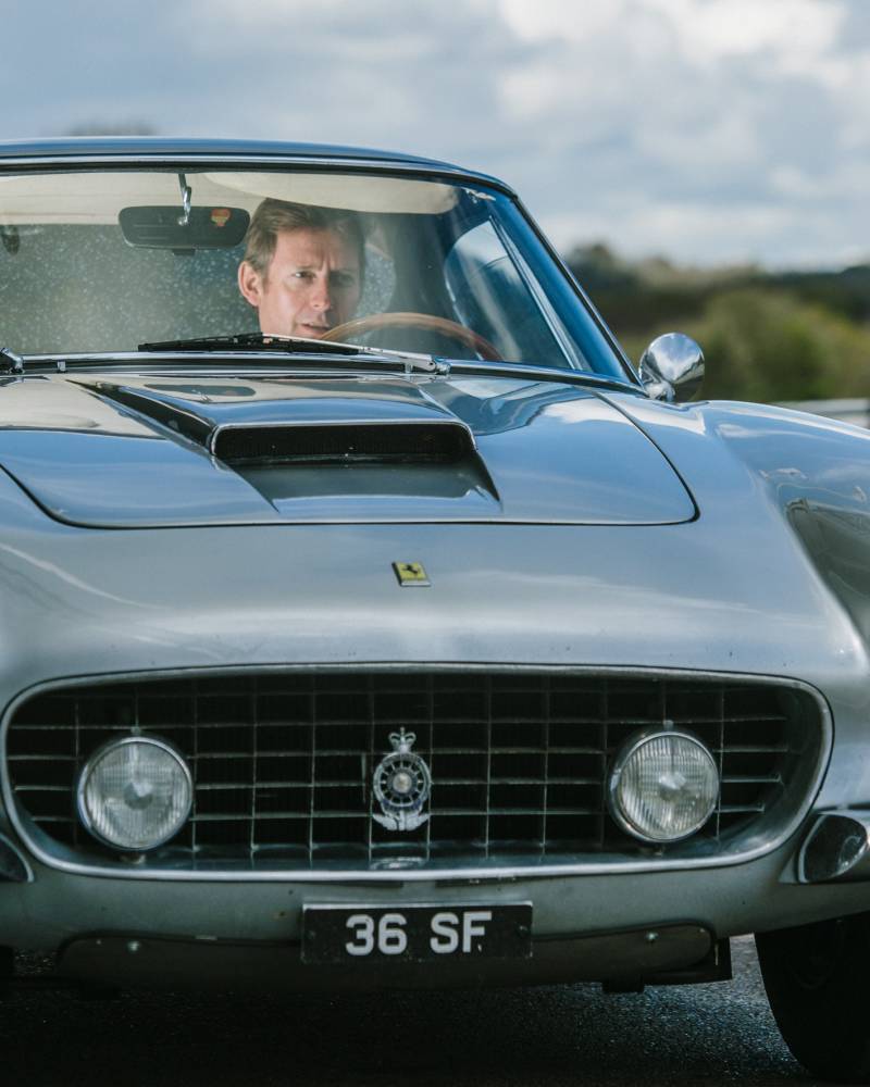 A man drives a classic silver Ferrari with a prominent hood scoop and a vintage license plate.