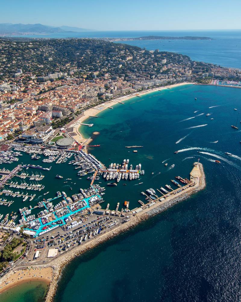 An aerial view captures the bustling marina and vibrant coastline of Cannes, France, dotted with numerous yachts under a clear blue sky.
