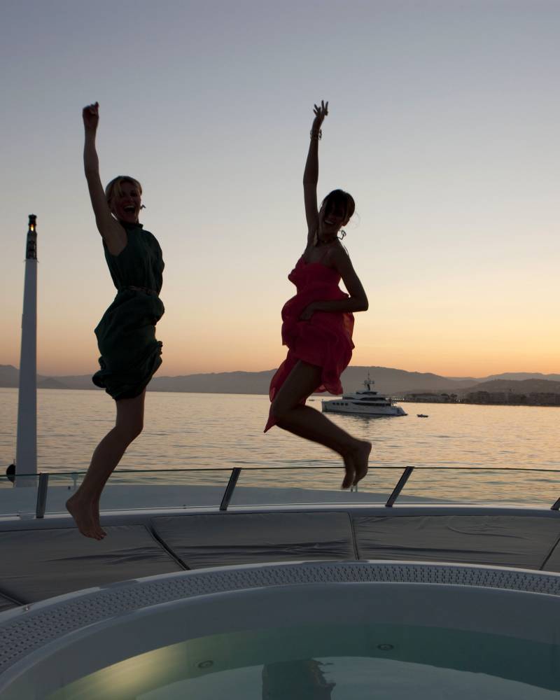 Two women in flowing dresses leap joyfully on a yacht deck at sunset with a scenic coastal backdrop.