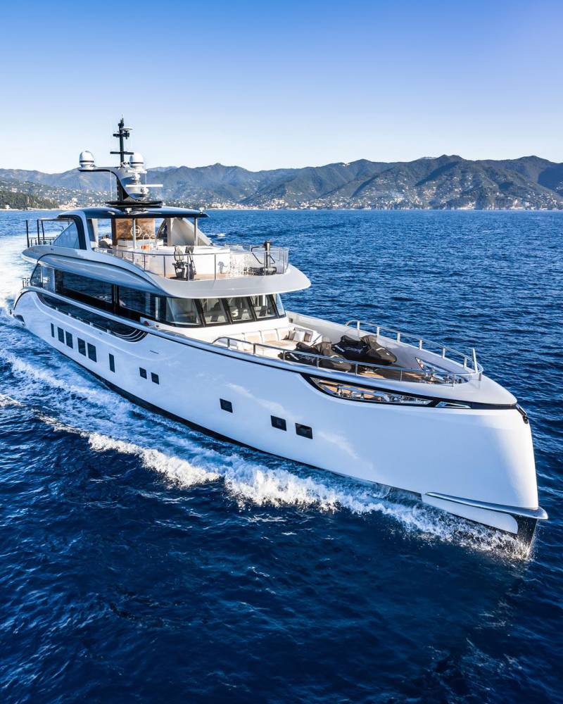 A sleek white yacht glides through the deep blue sea, with a picturesque coastline of rolling green hills in the background.