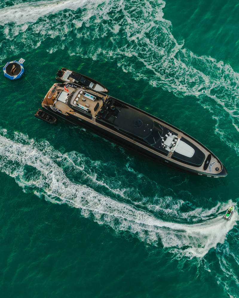 A sleek black yacht cruises through vibrant turquoise waters, accompanied by a jet ski and a floating water trampoline.