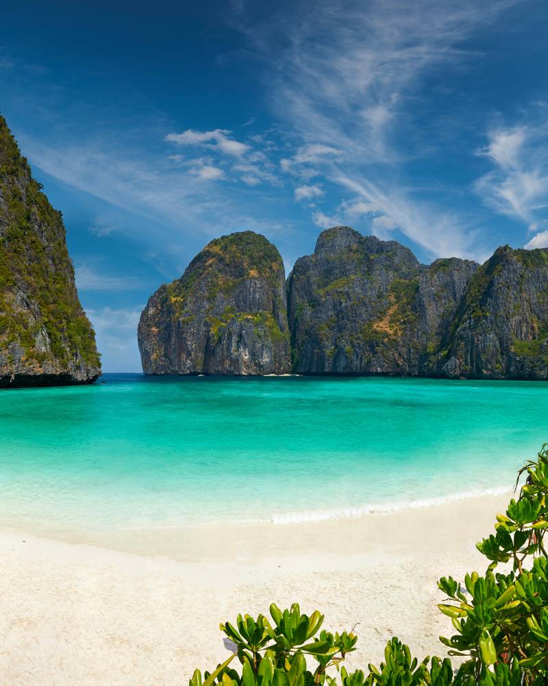 A stunning tropical beach with crystal-clear turquoise waters, surrounded by towering limestone cliffs and lush vegetation in the foreground.
