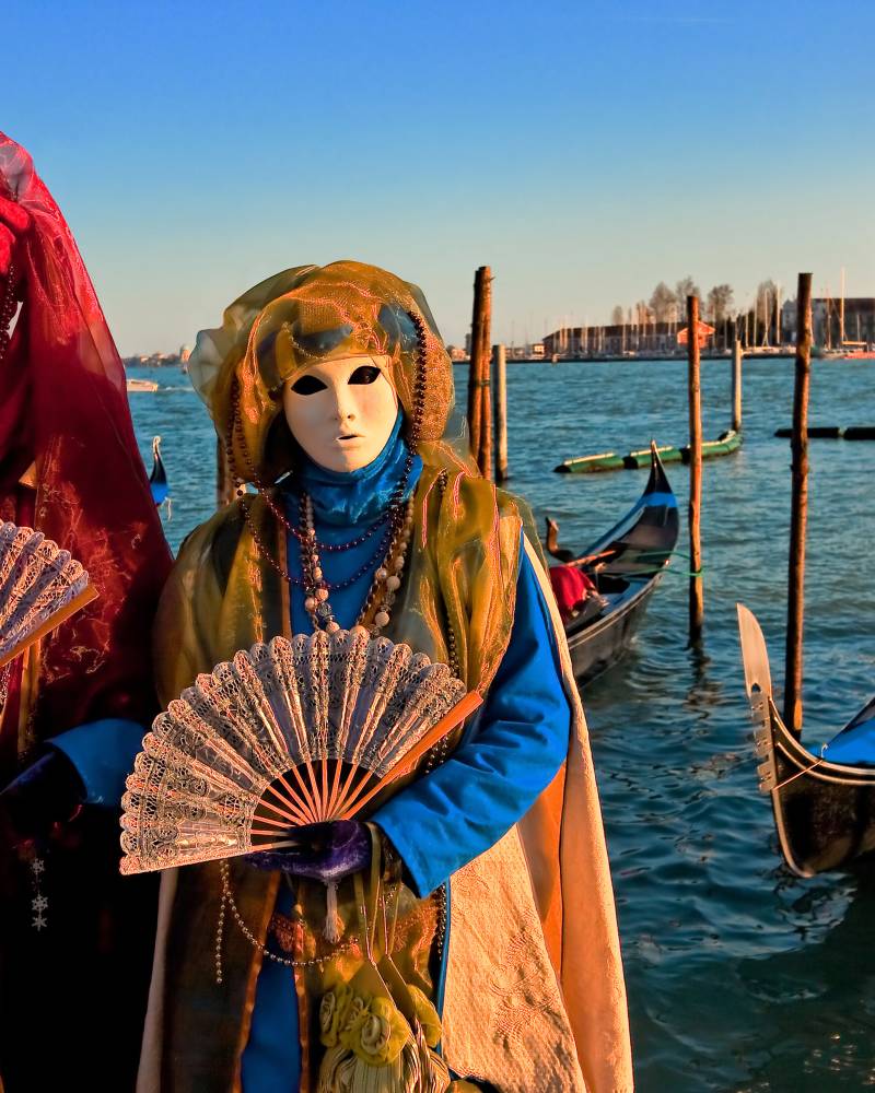 Venetian masquerade participants hold ornate fans against a backdrop of gondolas and historic architecture.
