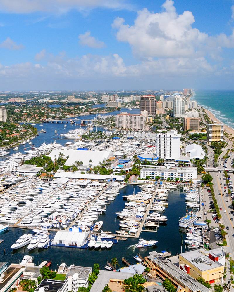 A bustling coastal city with a vibrant marina filled with boats, surrounded by high-rise buildings and lush greenery against a backdrop of a clear blue sky with fluffy white clouds.