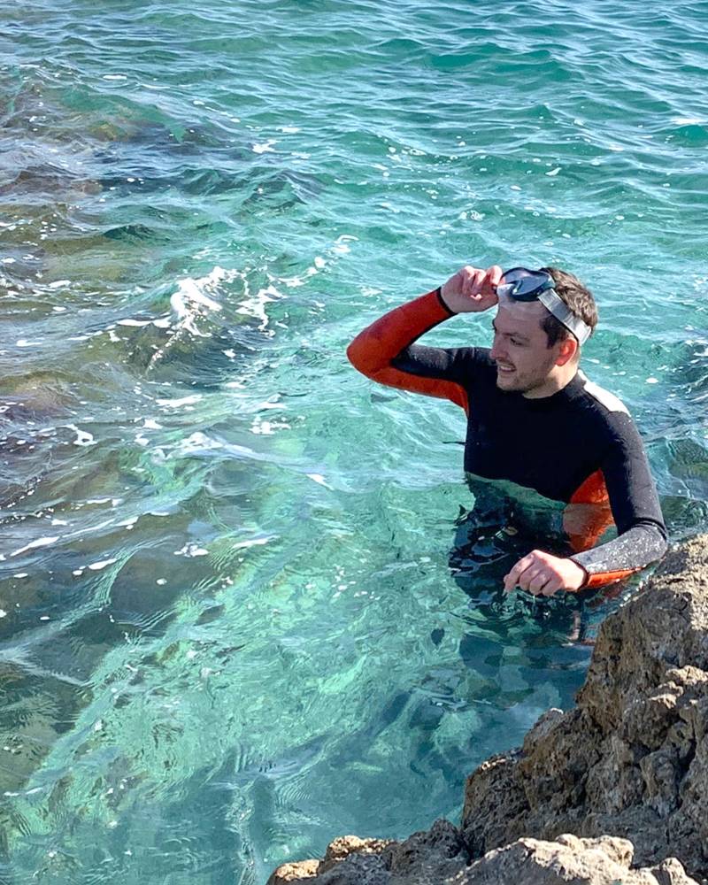 A man in a wetsuit adjusts his goggles while standing in the clear turquoise waters of a rocky coastline.