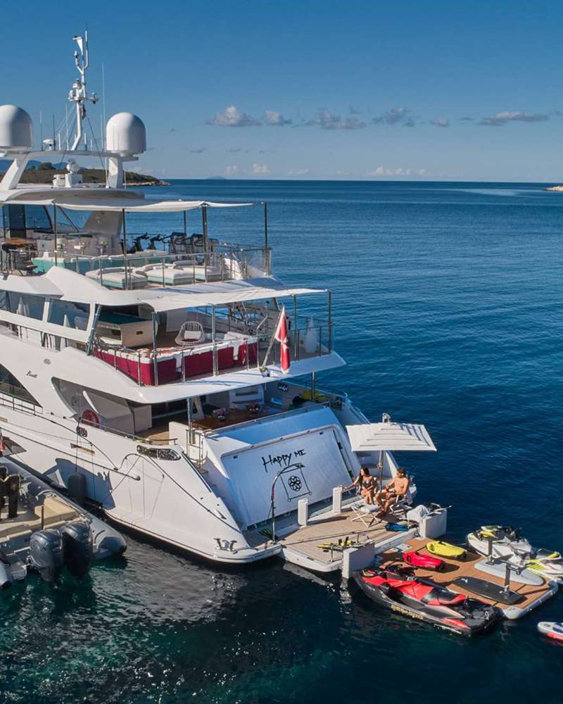 A luxurious yacht is anchored in a clear blue bay, surrounded by various watercraft and a distant sailboat.