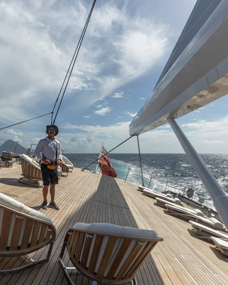 A man in a hat stands on the wooden deck of a yacht, holding a remote control as he surveys the open ocean.