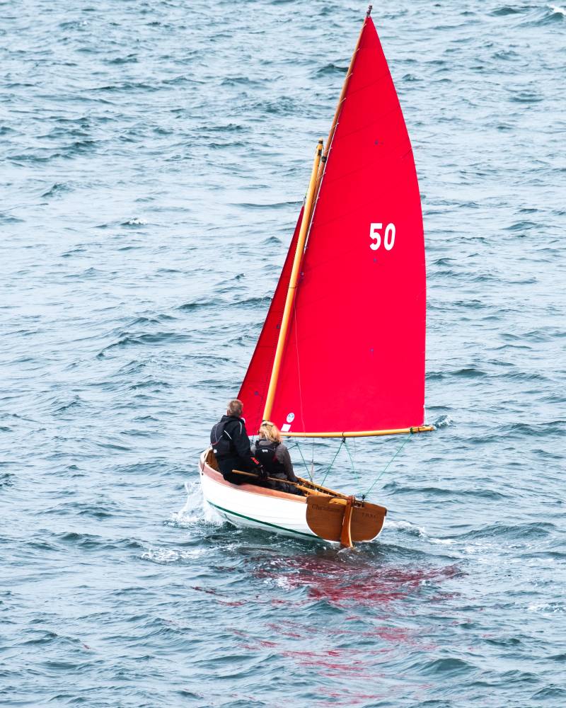 Two people sail a small boat with a bright red sail across the choppy blue water.