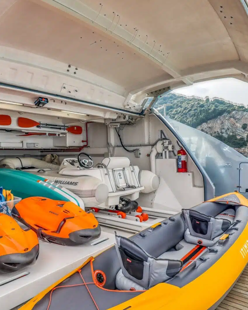 The image shows the interior of a boat or yacht, with various equipment and gear such as life jackets, bags, and other supplies neatly arranged on the deck. In the background, a scenic coastal landscape can be seen, suggesting the boat is situated in a picturesque location.