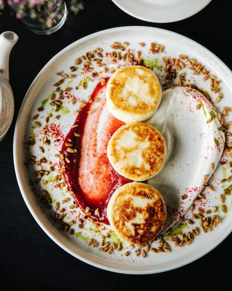 Golden-brown syrniki are artfully arranged with vibrant berry coulis and a dollop of cream, sprinkled with seeds and herbs.