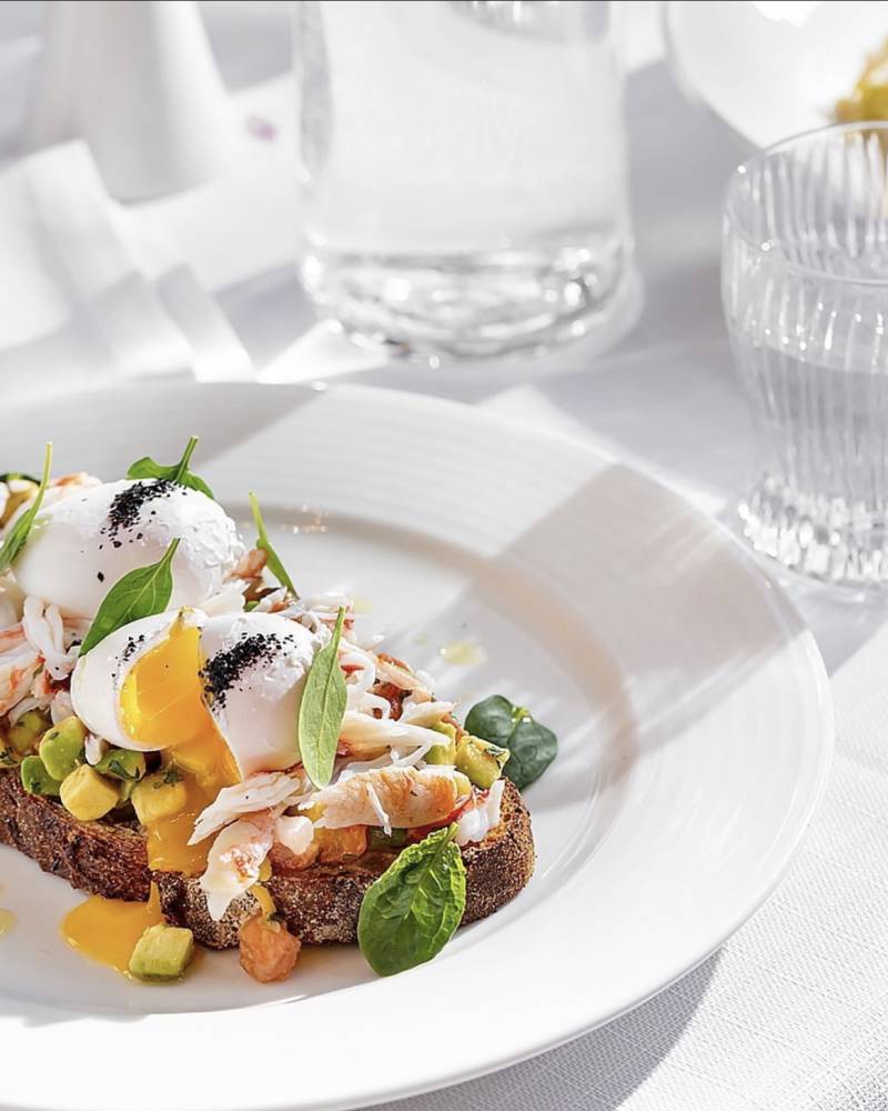 A perfectly poached egg crowns a decadent crab and avocado toast, drizzled with golden yolk on a crisp slice of dark bread.