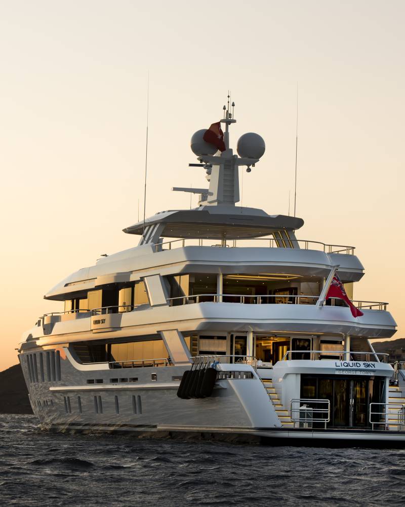 A luxurious yacht named Liquid Sky sails on calm waters under a warm, dusky sky near a coastal town.