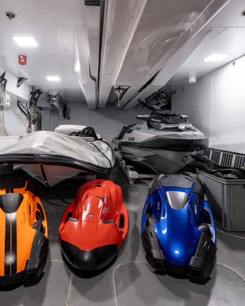 Three colorful personal watercraft are neatly stored alongside dive tanks and a jet ski inside a boat's spacious garage.