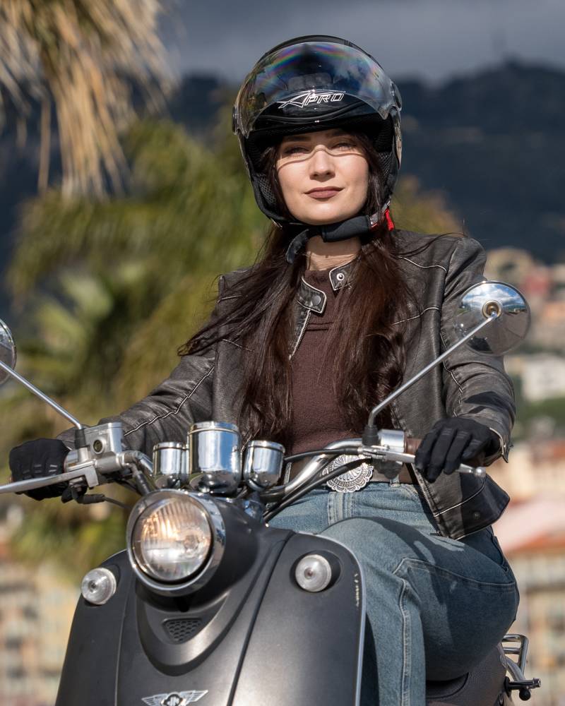 A stylish woman in a leather jacket and helmet rides a scooter through a sun-drenched coastal town.