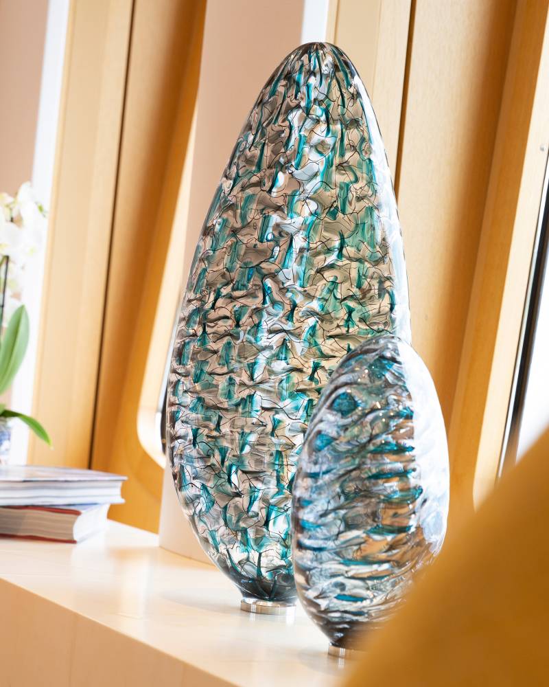 Two abstract glass sculptures with teal and silver patterns sit on a shelf next to a potted orchid.
