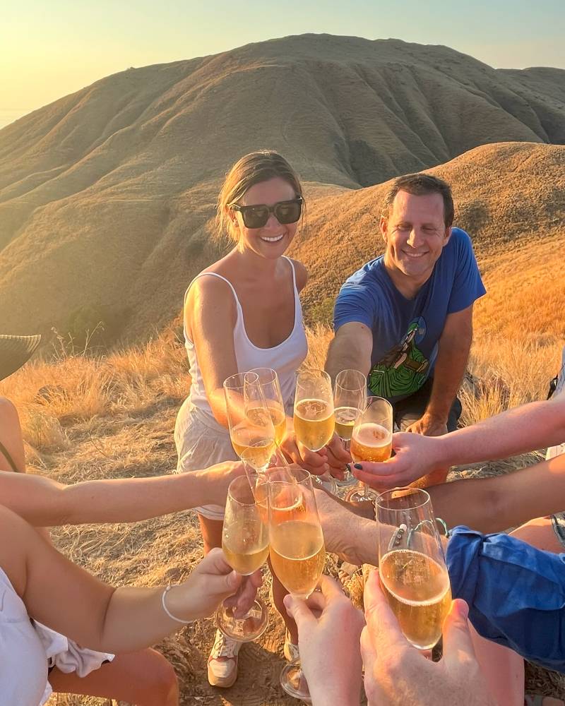 A group of people enjoying drinks together on a scenic mountain landscape with a warm, golden sunset in the background.