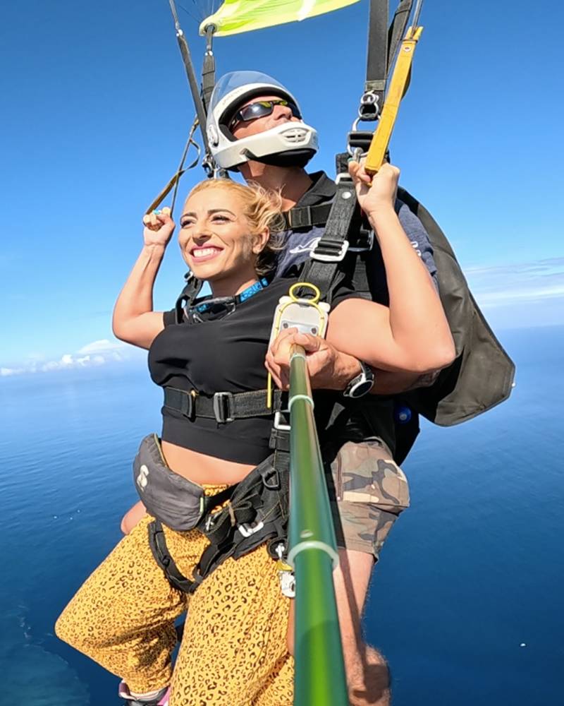 A smiling woman in leopard print pants enjoys a tandem skydive over a tropical ocean with a distant island.