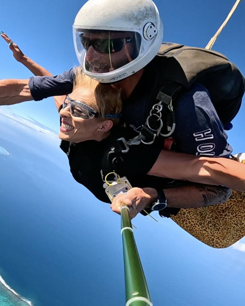 Two smiling skydivers give a thumbs-up as they freefall over a tropical coastline.