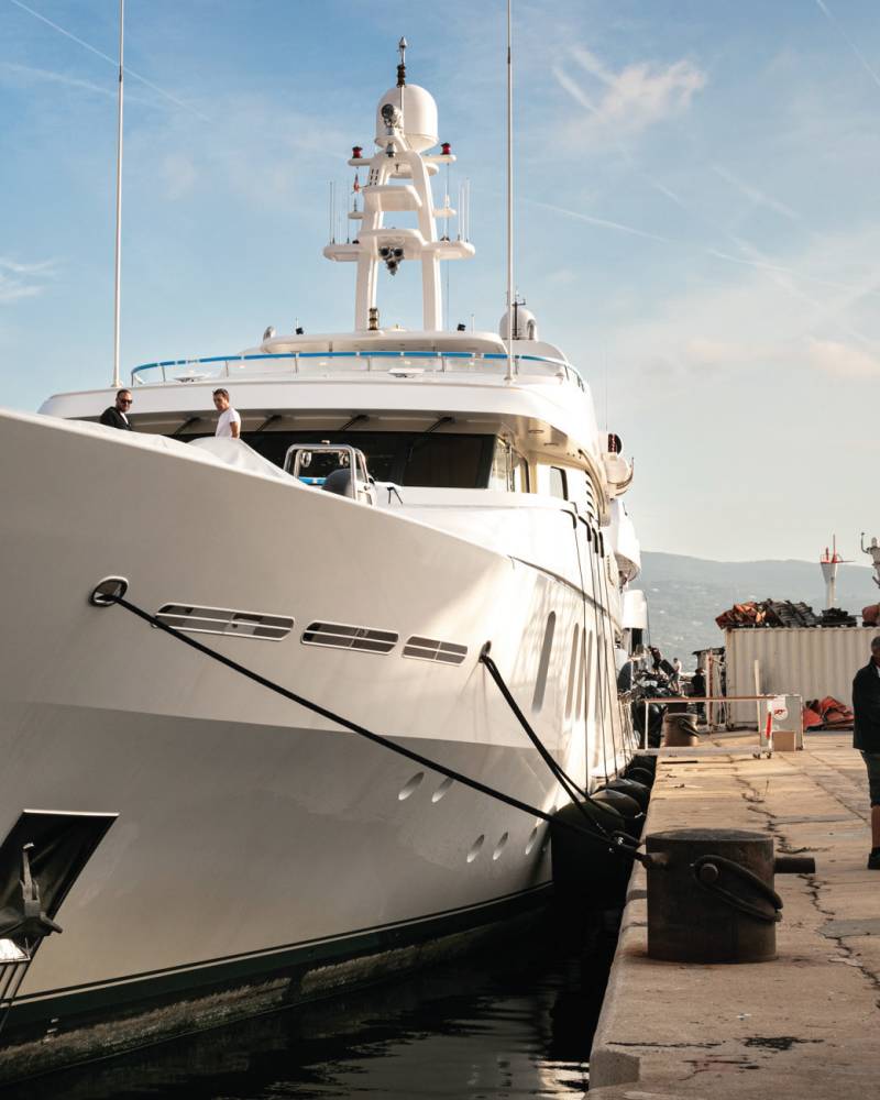 A massive white yacht is docked at a sunny harbor with a group of men gathered on the pier.