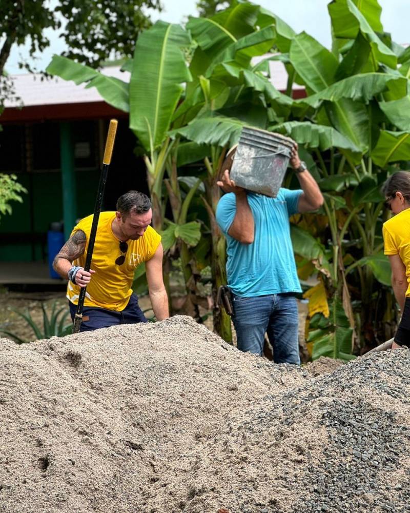 Volunteers in bright yellow shirts work together, shoveling sand and carrying buckets amidst lush banana trees.