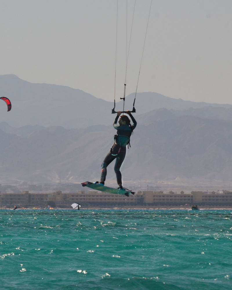 A kitesurfer soars through the air above turquoise waters with a hazy mountain range in the background.