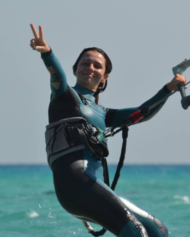 A kiteboarder smiles as she raises her hand in a peace sign while riding on the turquoise ocean.
