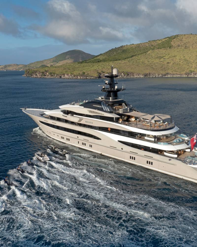 A luxurious superyacht cruises through the deep blue sea, trailed by a fleet of jet skis and tenders near a lush, green coastline.