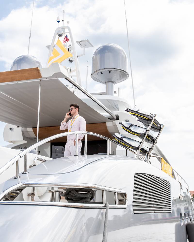 A man in a pink shirt talks on his phone while standing on the deck of a luxurious yacht.