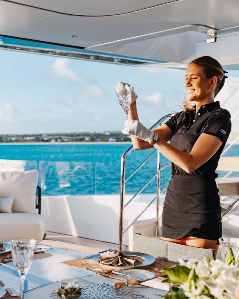 A smiling stewardess polishes a glass on a yacht deck set for a meal overlooking the turquoise ocean.