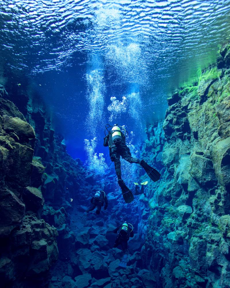 Divers explore the crystal-clear waters of a submerged fissure, surrounded by rugged rock formations and rising bubbles.