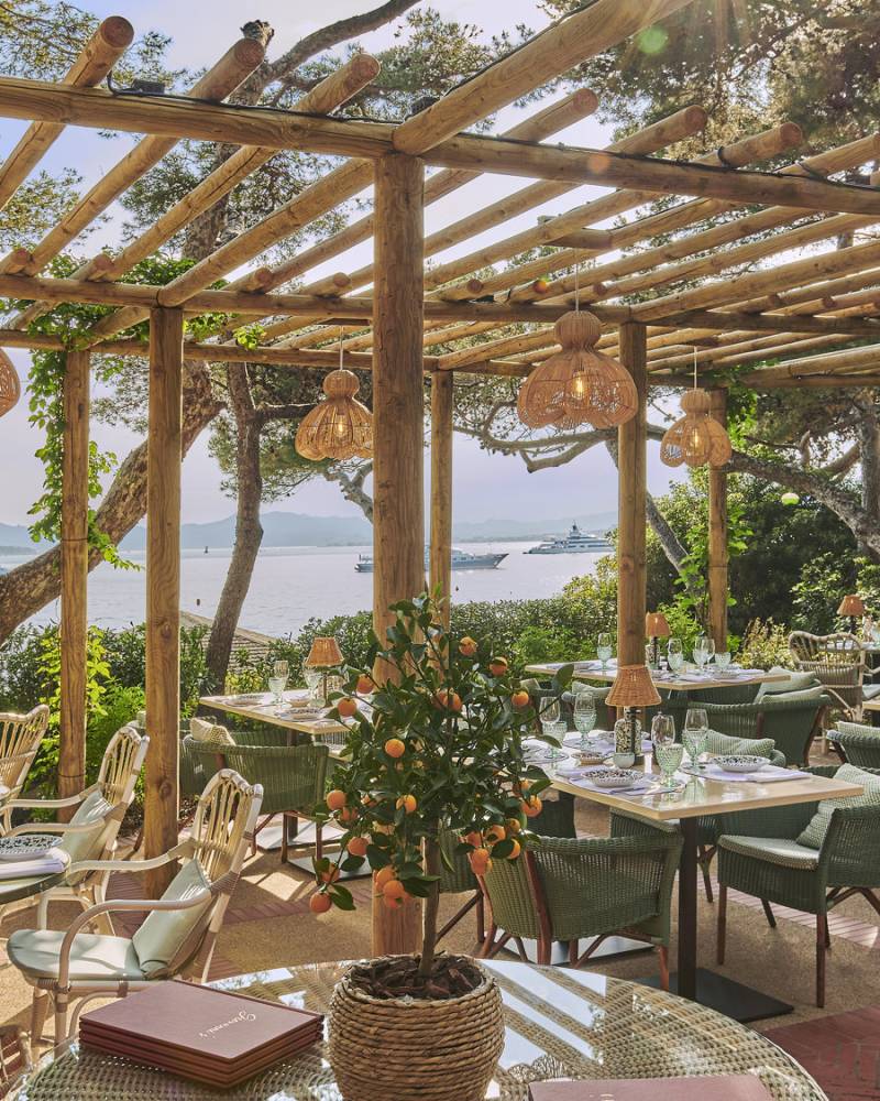 Dine al fresco under a wooden pergola overlooking the sparkling sea, surrounded by lush greenery and elegant table settings.