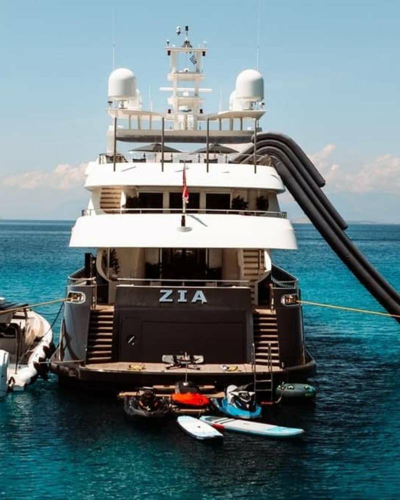 A luxurious yacht named Zia is anchored in crystal-clear turquoise waters, featuring an inflatable slide and various watercraft.