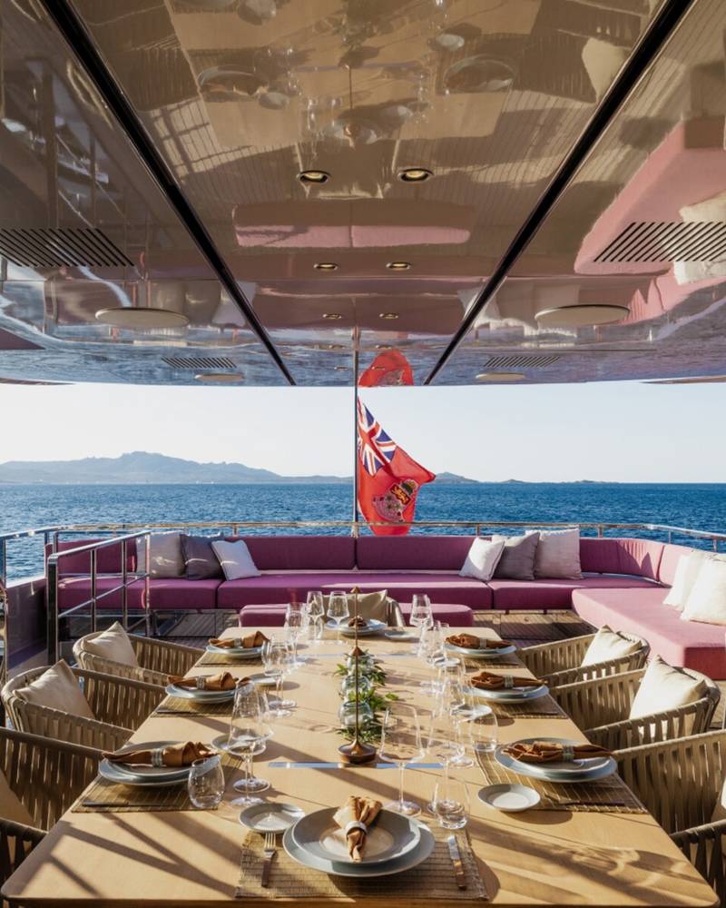 A long dining table is set for a meal on the deck of a luxury yacht, overlooking the sparkling blue sea.