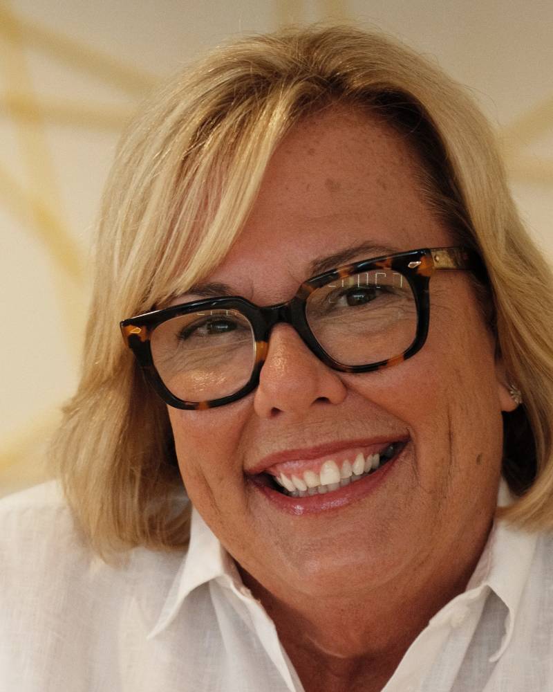 A woman with blonde hair and tortoiseshell glasses smiles warmly, her white shirt catching the light.