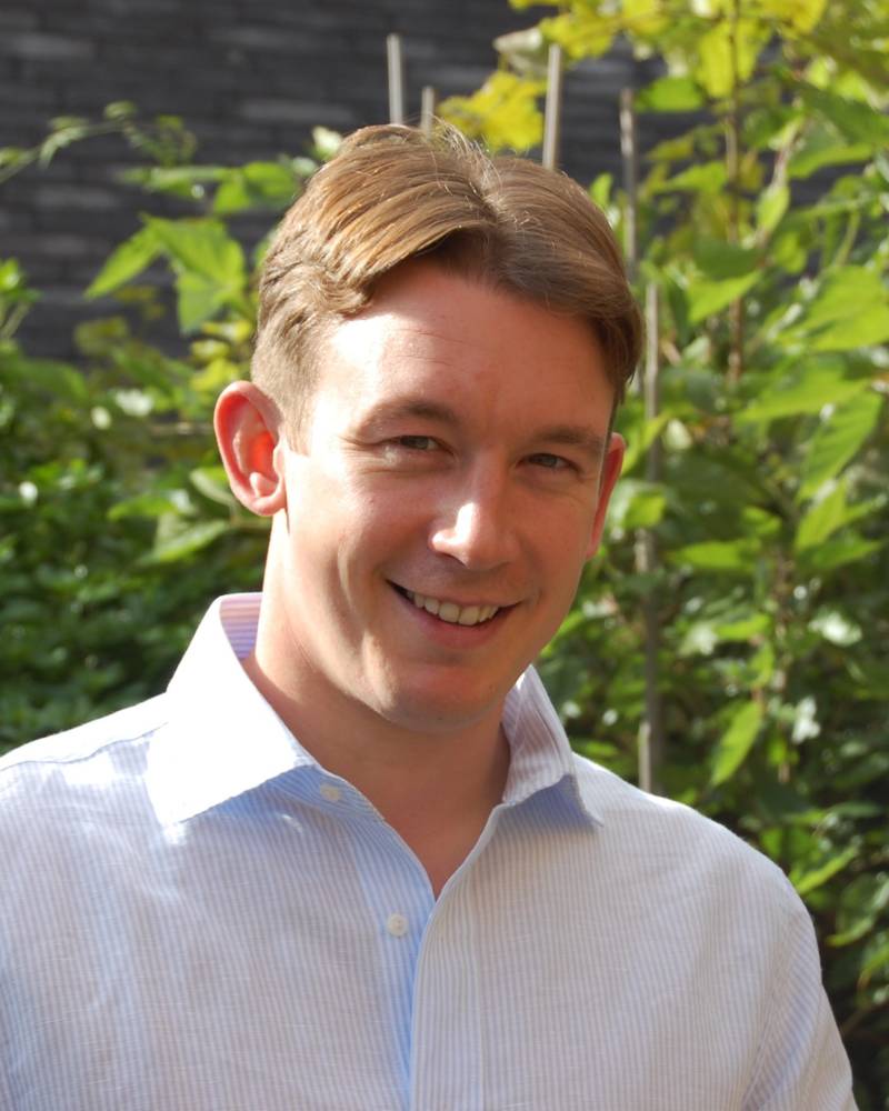 A man smiles warmly while posing outdoors amidst lush green foliage and a brick wall.